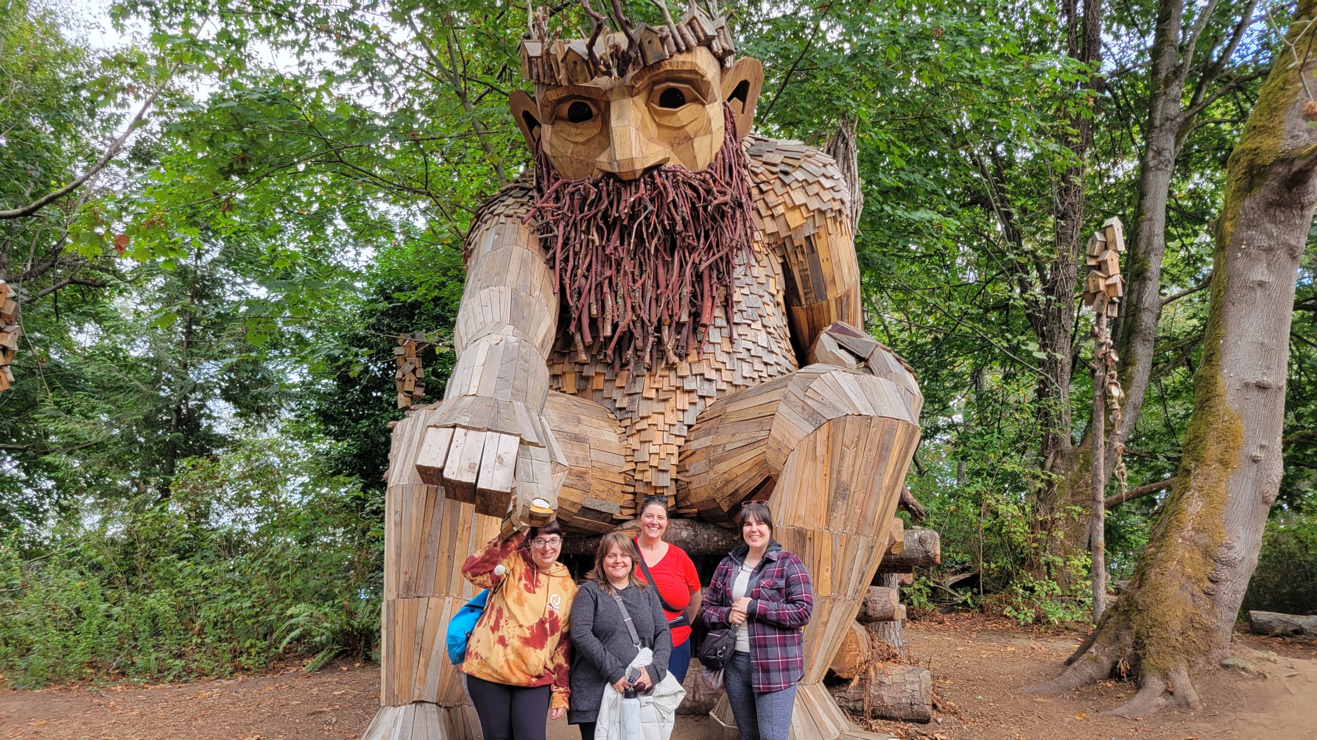 a group of friends stand at the feet of a wood troll, posing for a picture.