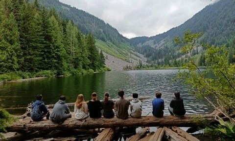 a group of campers takes on the view while sitting on a log next to a lake