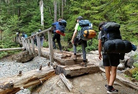 a group of campers hiking with full bags on their backs as they cross over a bridge