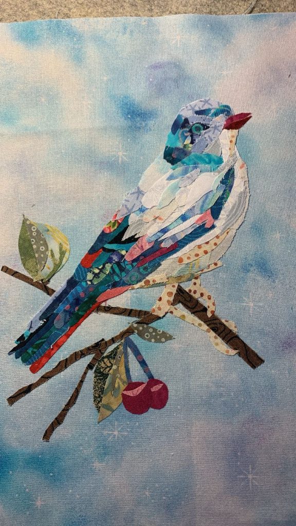 Collage Quilting of a bird on a small branch with a mottled blue background