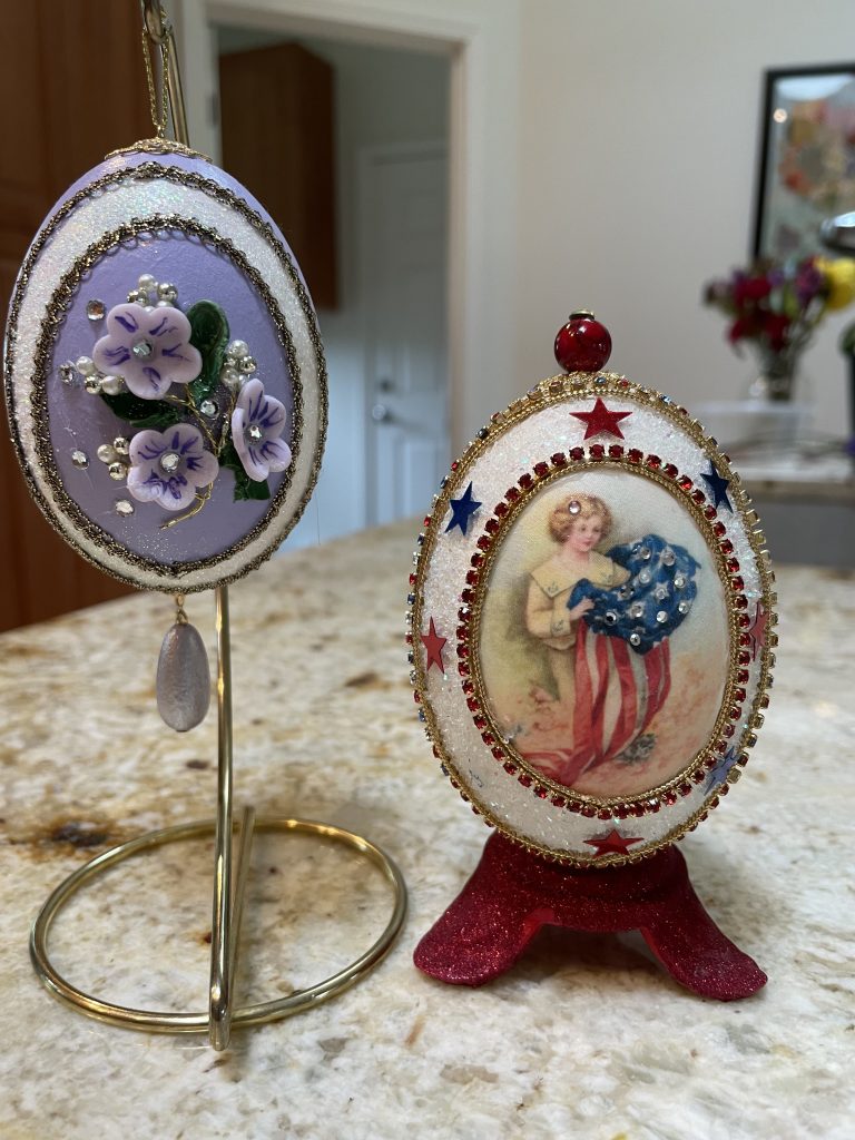 2 Decorated eggs. One is lavender and white with small flowers and it is hanging on a display hook. The other is decorated in patriotic colors with an illustration of a woman holding a US flag and it is displayed on a small stand.
