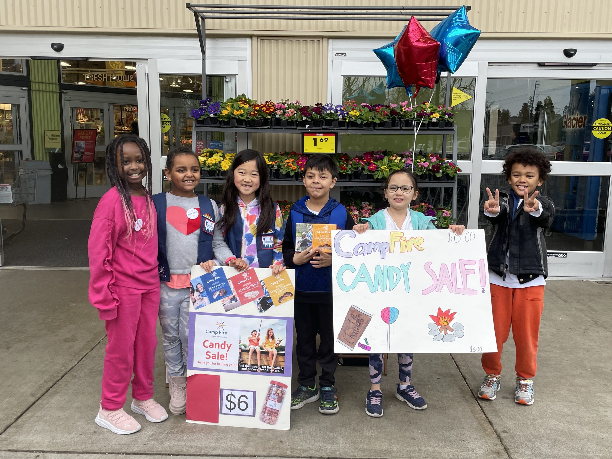Six youth standing in front of a store display of flowers with red and blue baloons holding hand-made candy sale signs at their site sale