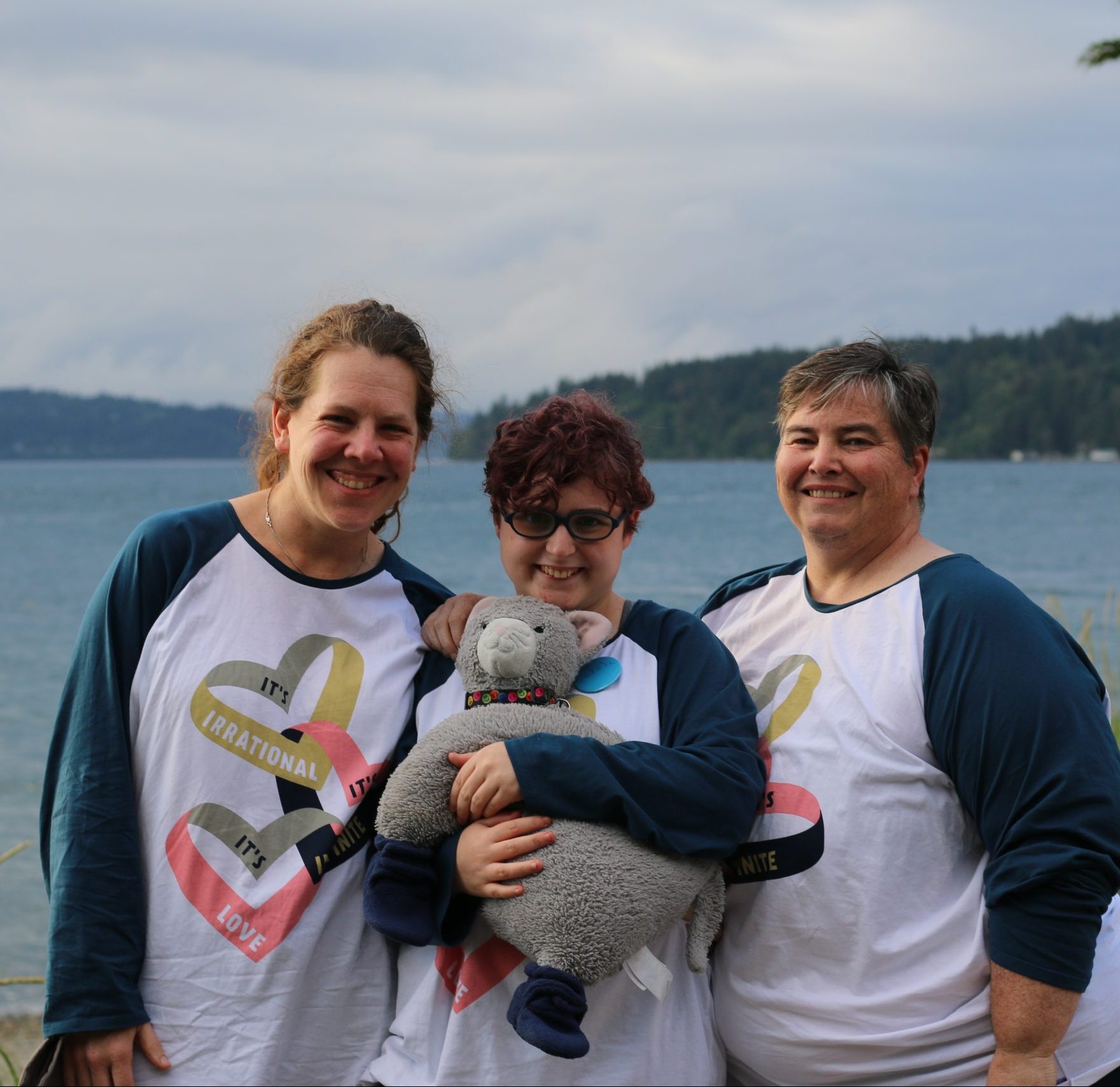 A family of three stand wearing matching shirts with hearts in front of the Puget Sound at Camp Sealth