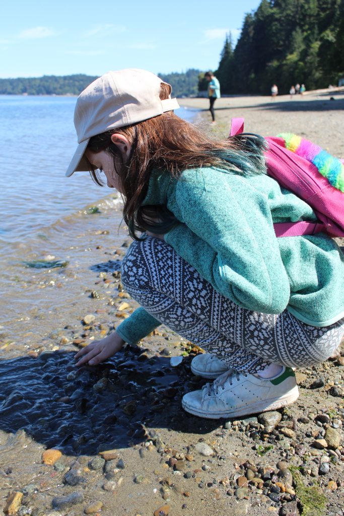 Student in blue jacket and pink backpack squats on the beach to look at rocks and shells near the waters edge