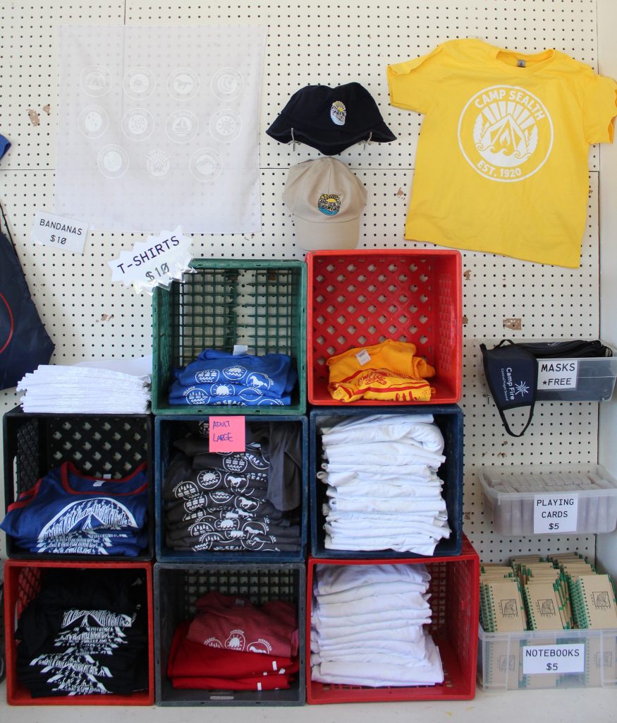 Milk crates holding t-shirts and items like bandanas, notebooks, and t-shirts on display on the wall behind at the Camp Store
