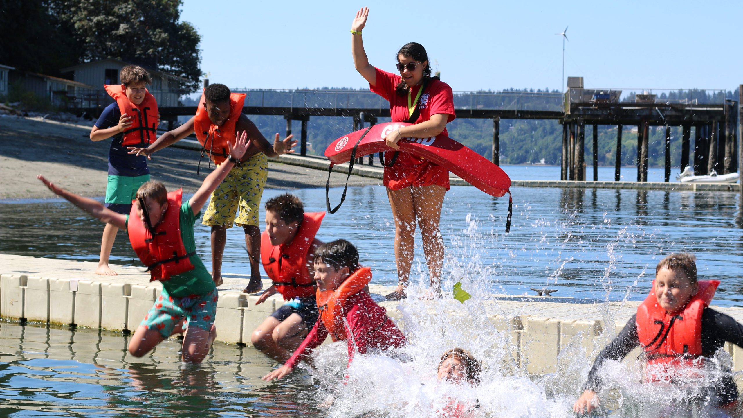 Campers in orange life vests splash into the Puget Sound with a lifeguard standing on the dock
