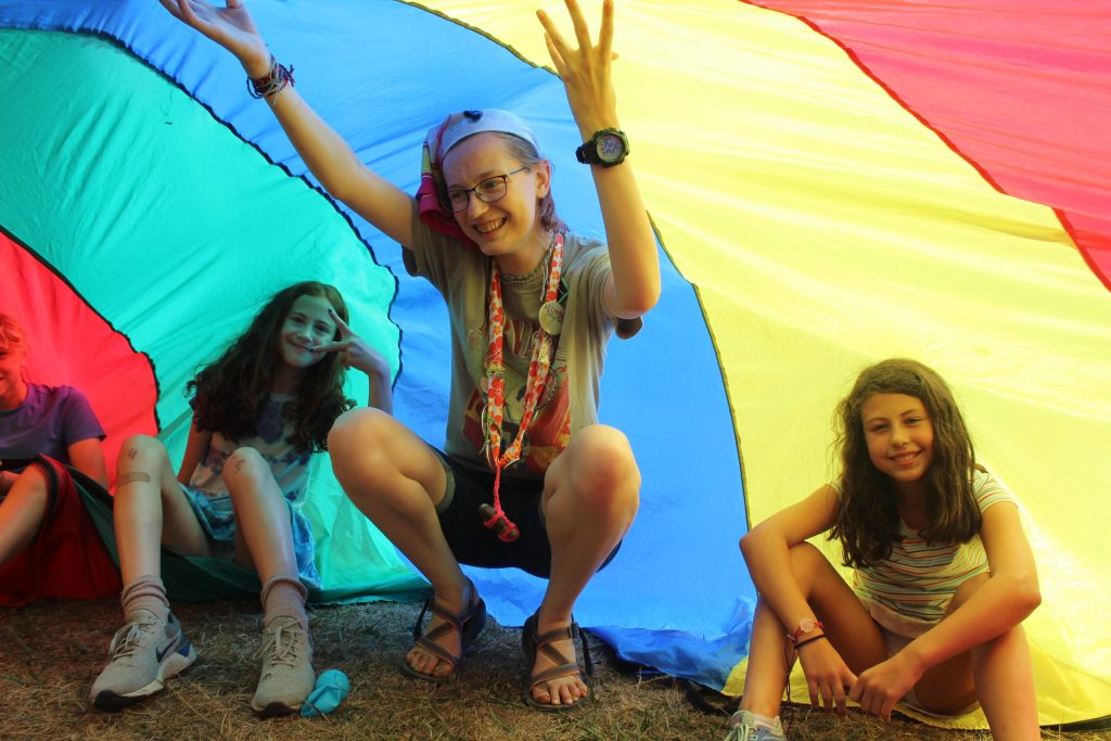 A staff member and campers play underneath a colorful rainbow parachute