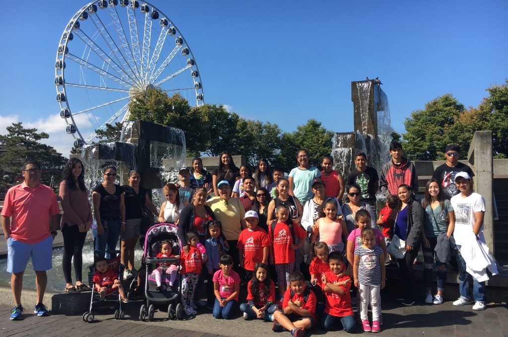 A group of families stands together for a photo with the Great Seattle Wheel behind. Some family members are wearing red shirts with a white Camp Fire logo.