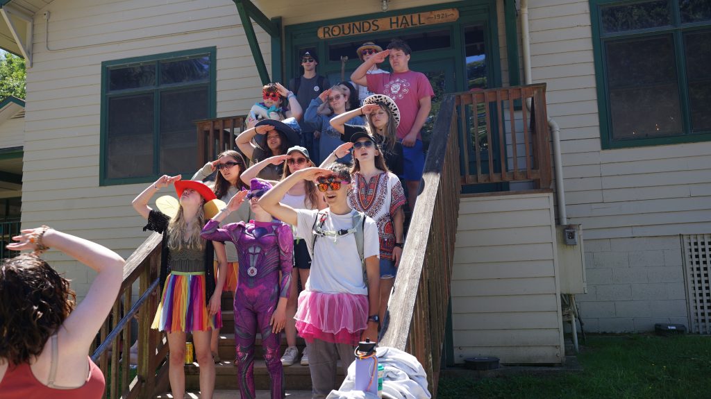 a group of campers dressed in costume and saluting the camp leader while posing on a staircase.