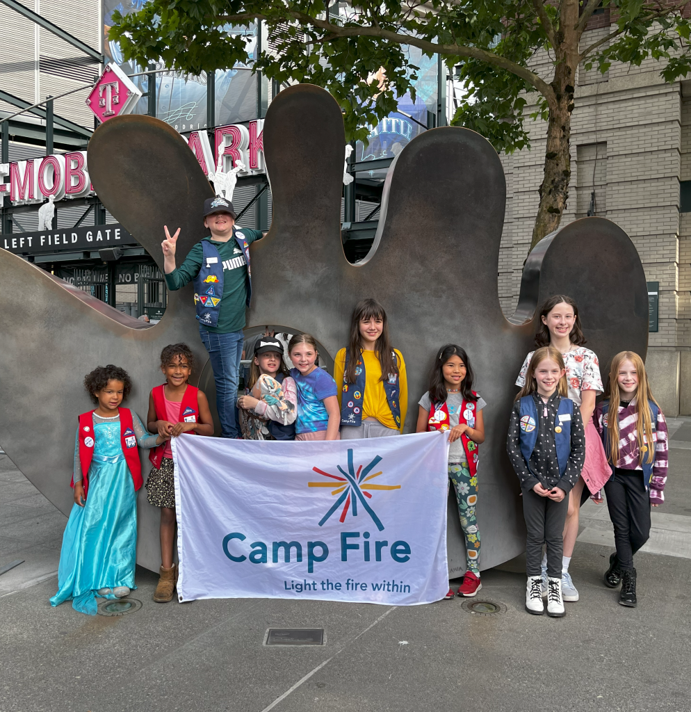 Camp Fire youth stand with a white flag featuring the Camp Fire logo in front of a baseball glove statue at T-Mobile Park